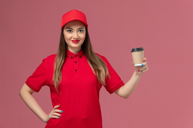 Front view female courier in red uniform and cape holding delivery coffee cup on the pink background service delivery uniform job worker