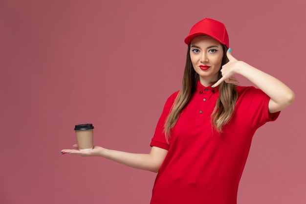 Front view female courier in red uniform and cape holding delivery coffee cup on pink background service delivery uniform job