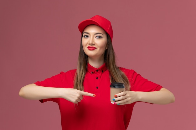 Front view female courier in red uniform and cape holding delivery coffee cup on the pink background service delivery uniform job