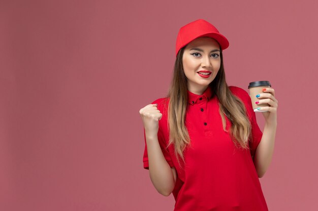 Вид спереди женщина-курьер в красной форме и накидке, держащая чашку кофе для доставки и радующаяся на розовом фоне, униформа службы доставки, рабочий