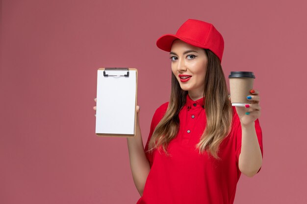 Front view female courier in red uniform and cape holding delivery coffee cup with notepad on pink background service delivery uniform