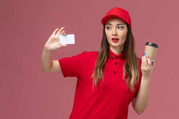 Front view female courier in red uniform and cape holding delivery coffee cup with white card on pink background service job delivery worker uniform