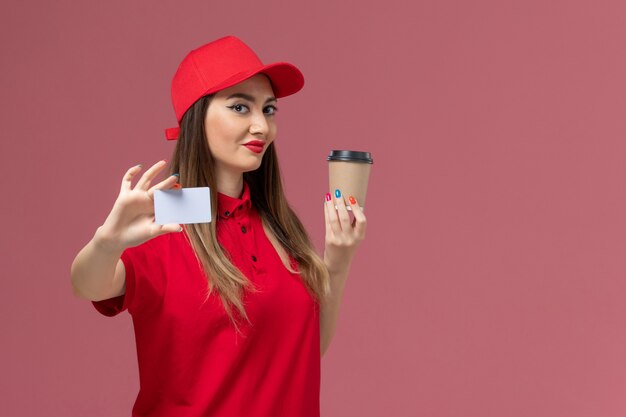 Front view female courier in red uniform and cape holding delivery coffee cup with white card smiling on pink background service job delivery worker uniform