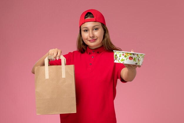 Front view female courier in red uniform and cape holding delivery food package and bowl on pink wall, uniform delivery job service