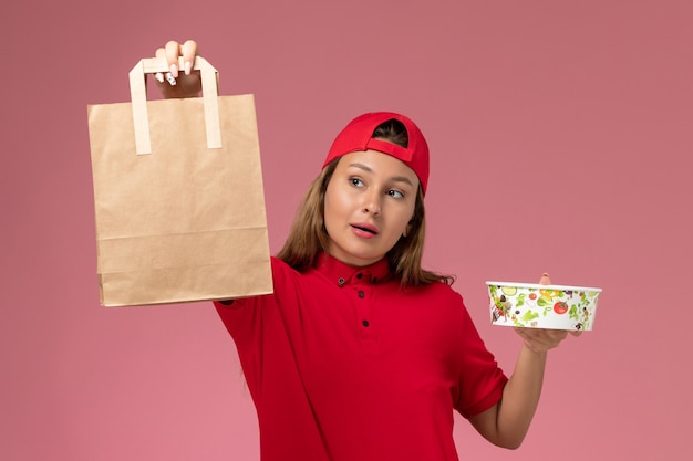 Free photo front view female courier in red uniform and cape holding delivery food package and bowl on the pink wall, uniform delivery job service