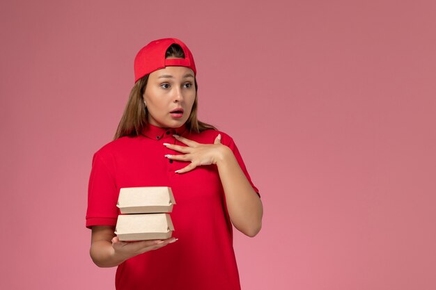 Front view female courier in red uniform and cape holding delivery food package on light-pink background uniform delivery service company job work