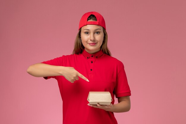 Вид спереди женщина-курьер в красной форме и накидке с доставкой продуктового пакета на светло-розовом фоне