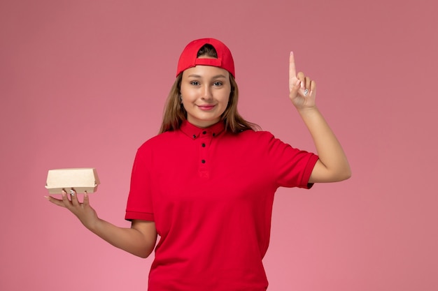 Front view female courier in red uniform and cape holding delivery food package on light-pink background worker uniform delivery service company job