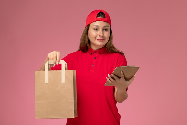 赤い制服を着た正面図の女性宅配便と淡いピンクの壁に配達食品パッケージとメモ帳を保持している岬、制服配達の仕事のサービス作業