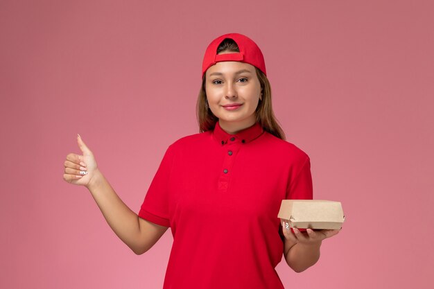 Front view female courier in red uniform and cape holding delivery food package on the pink background uniform delivery company job worker girl