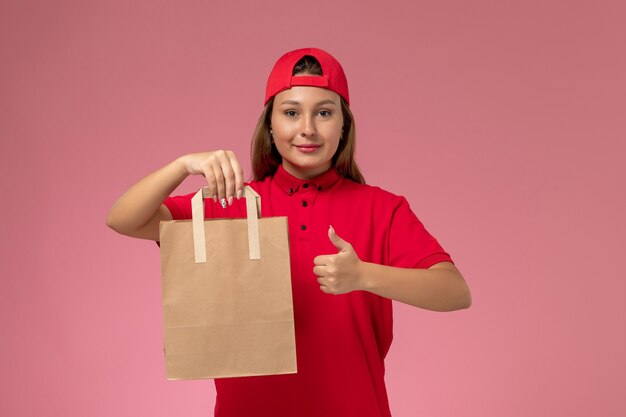 Front view female courier in red uniform and cape holding delivery food package on the pink background uniform delivery job service work
