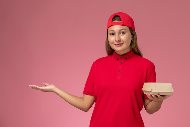 Front view female courier in red uniform and cape holding delivery food package on the pink background uniform delivery service company girl