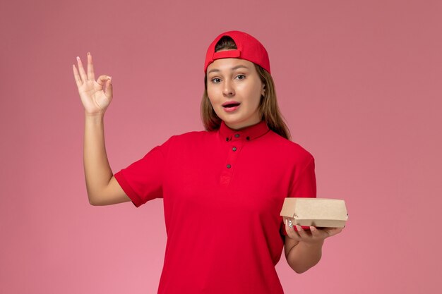 Front view female courier in red uniform and cape holding delivery food package on the pink background uniform delivery service