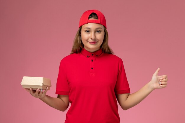 Front view female courier in red uniform and cape holding delivery food package on pink background uniform delivery worker service company job