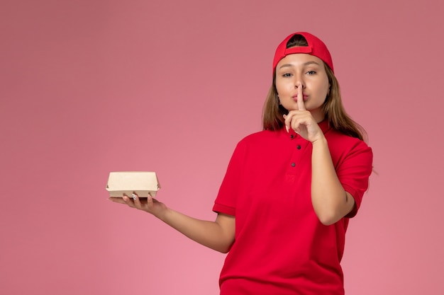Front view female courier in red uniform and cape holding delivery food package on pink background worker uniform delivery service company job work