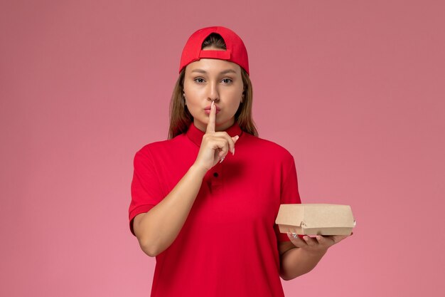 Front view female courier in red uniform and cape holding delivery food package on the pink wall, uniform delivery service company job worker