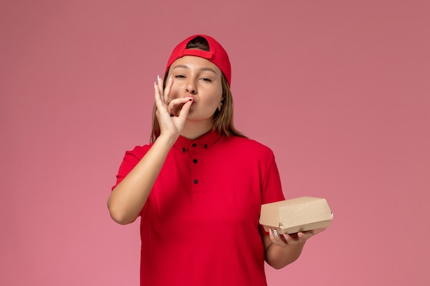 Front view female courier in red uniform and cape holding delivery food package on the pink wall, uniform delivery service company work girl