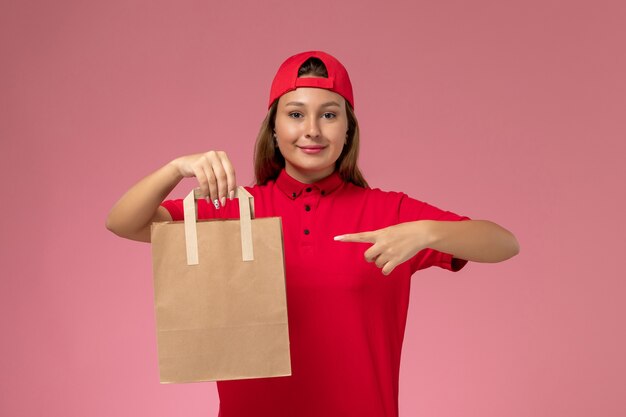 赤い制服を着た正面図の女性の宅配便とピンクの壁に微笑んで配達食品パッケージを保持している岬、制服配達ジョブサービス