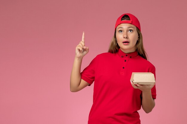 Front view female courier in red uniform and cape holding delivery food package and thinking on pink wall, uniform delivery service company job worker