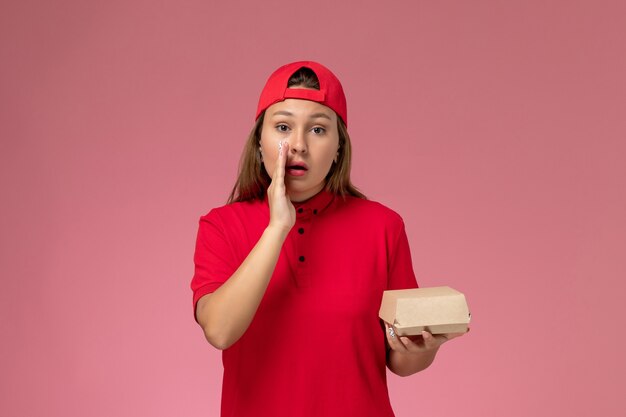 Front view female courier in red uniform and cape holding delivery food package whispering on the pink wall, uniform delivery service company job