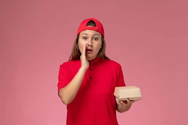 Front view female courier in red uniform and cape holding delivery food package whispering on the pink wall, uniform delivery service company job
