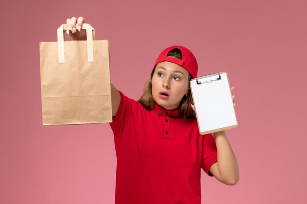 Front view female courier in red uniform and cape holding delivery food package with notepad on light pink wall, uniform delivery job service worker