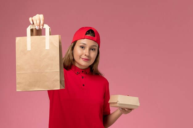 Front view female courier in red uniform and cape holding delivery food packages on the pink wall, uniform delivery service