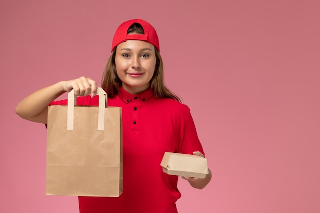 Corriere femminile di vista frontale in uniforme rossa e mantello che tiene i pacchetti di cibo di carta di consegna sulla parete rosa, servizio di consegna uniforme