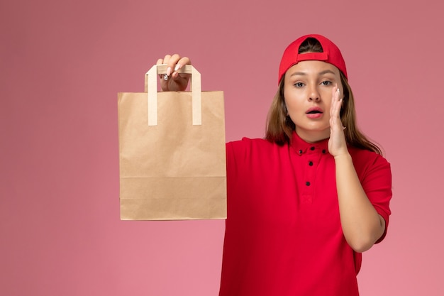 Front view female courier in red uniform and cape holding delivery paper package on pink wall, job uniform delivery service job