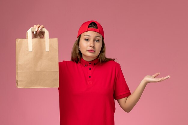 Женщина-курьер в красной форме и накидке, держащая бумажный пакет для доставки на розовой стене, вид спереди, работа службы доставки униформы