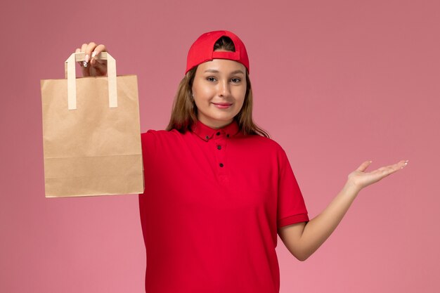Front view female courier in red uniform and cape holding delivery paper package on pink wall, job uniform delivery service worker