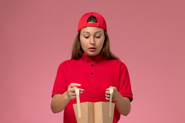 Front view female courier in red uniform and cape holding delivery paper package on pink wall, job uniform delivery service