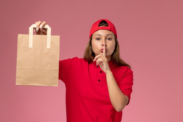 Front view female courier in red uniform and cape holding delivery paper package on pink wall, job uniform delivery work service