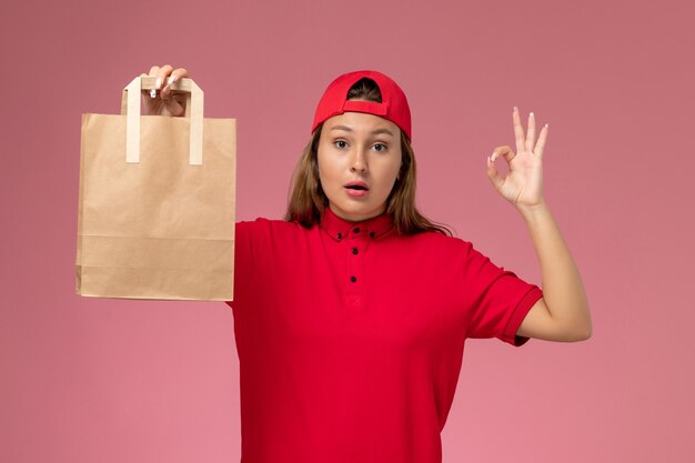 Front view female courier in red uniform and cape holding delivery paper package on the pink wall, uniform delivery service