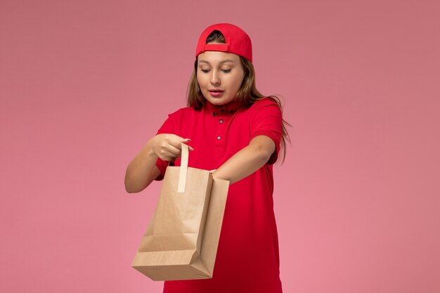 front view female courier in red uniform and cape holding delivery paper package taking something out of it on pink wall, uniform delivery service
