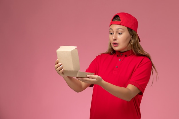 Front view female courier in red uniform and cape holding empty delivery food package on light-pink wall, uniform delivery service company job