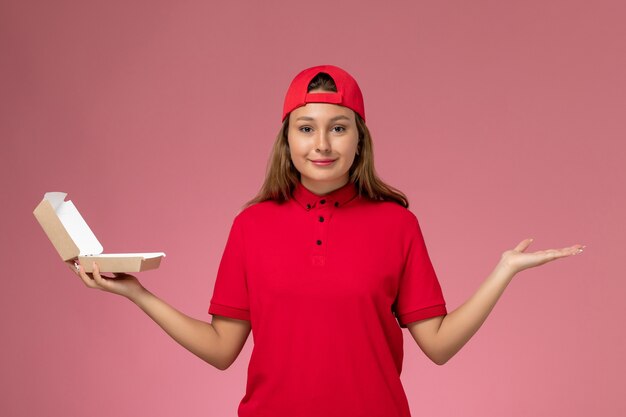 Front view female courier in red uniform and cape holding empty delivery food package on light-pink wall, uniform delivery service company worker