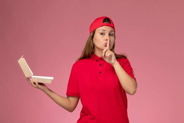 Corriere femminile di vista frontale in uniforme rossa e mantello che tiene il pacchetto di cibo di consegna vuoto sulla parete rosa chiaro, società di servizi di consegna uniforme