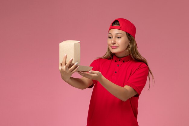 Front view female courier in red uniform and cape holding empty delivery food package opening it on light-pink wall, uniform delivery service company