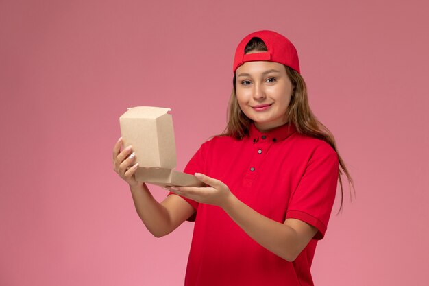 Front view female courier in red uniform and cape holding empty delivery food package and smiling on light-pink wall, uniform delivery service company