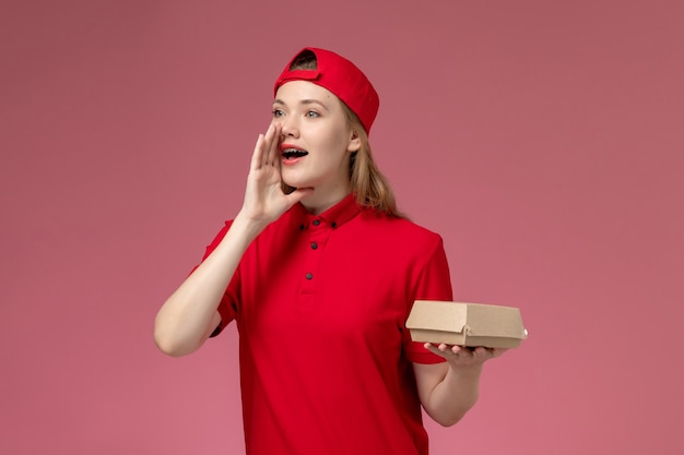 Front view female courier in red uniform and cape holding little delivery food package calling out on the pink wall, delivery job service company uniform