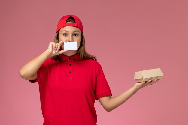 빨간 유니폼과 밝은 분홍색 벽에 작은 배달 음식 패키지와 카드를 들고 케이프, 균일 한 작업 노동자 배달 서비스에 전면보기 여성 택배