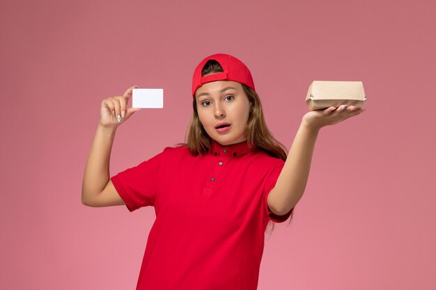 赤い制服とケープの正面図の女性の宅配便は、淡いピンクの壁に小さな配達食品パッケージとカードを保持し、制服の労働者配達サービス