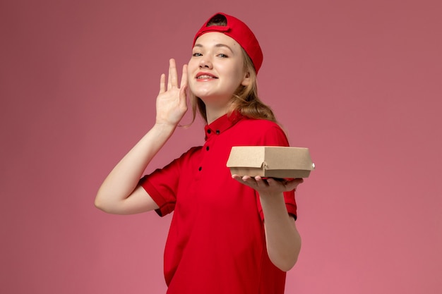 Вид спереди женщина-курьер в красной форме и накидке, держащая небольшой пакет с доставкой еды на светло-розовой стене, униформа компании службы доставки