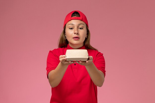 Front view female courier in red uniform and cape holding little delivery food package on light-pink wall, uniform delivery service company work