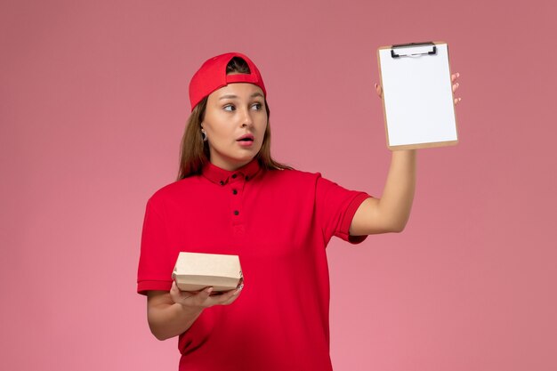 赤い制服とピンクの壁に小さな配達食品パッケージとメモ帳を保持している岬の正面図の女性の宅配便、制服配達サービス会社の労働者