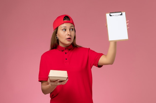 Front view female courier in red uniform and cape holding little delivery food package and notepad on pink wall, uniform delivery service company worker