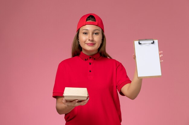 Front view female courier in red uniform and cape holding little delivery food package and notepad on pink wall, uniform delivery service company