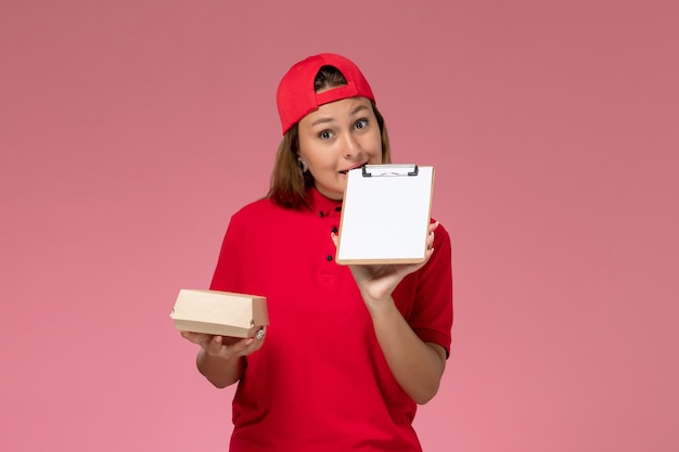 Front view female courier in red uniform and cape holding little delivery food package and notepad on the pink wall, uniform delivery service company