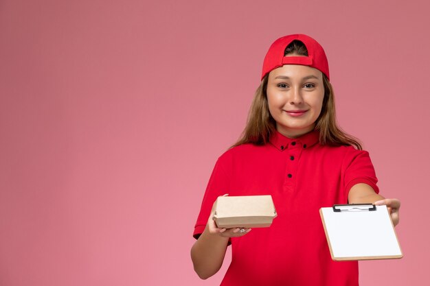 Free photo front view female courier in red uniform and cape holding little delivery food package and notepad smiling on the pink wall, uniform worker delivery service